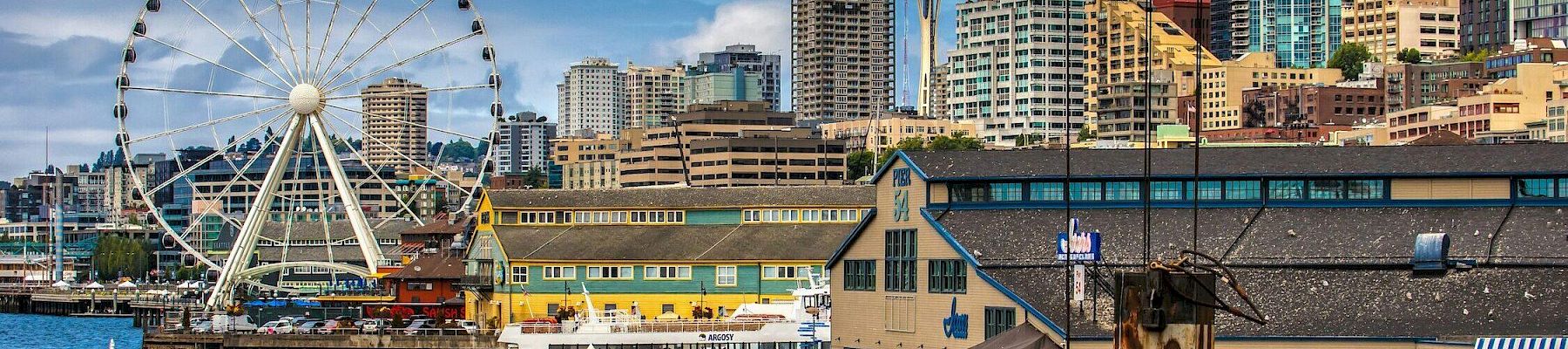 Seattle-style waterfront with a large Ferris wheel, skyline, and piers; colorful buildings and boats along the harbor.
