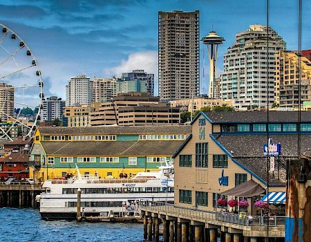 Seattle-style waterfront with a large Ferris wheel, skyline, and piers; colorful buildings and boats along the harbor.