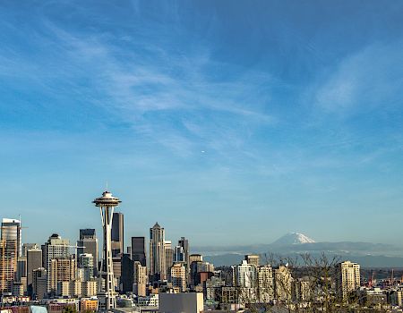 Seattle skyline with iconic Space Needle, numerous buildings, clear blue sky, distant snow-capped mountain, maybe Mount Rainier, sunny.