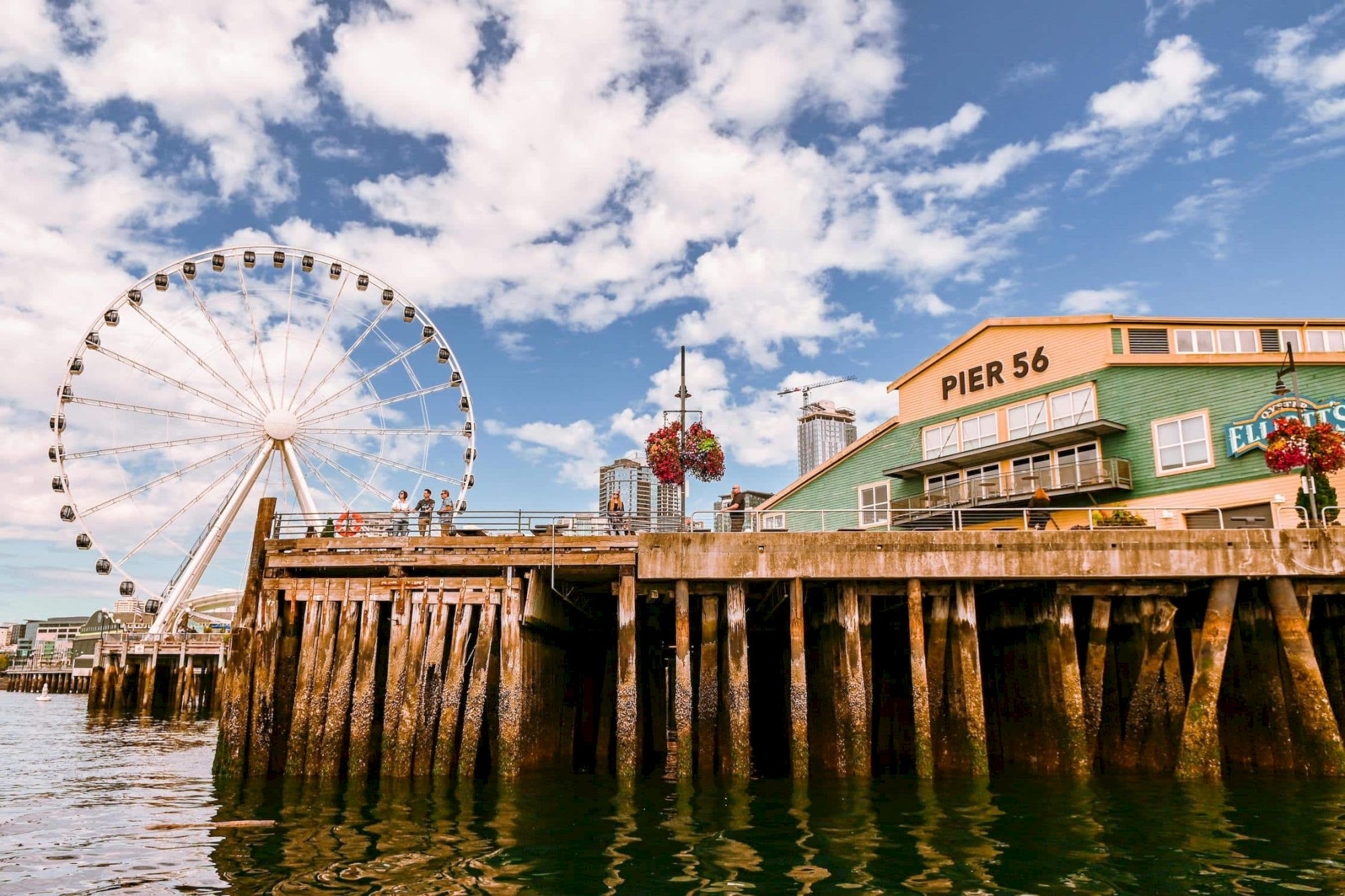 A seaside pier with sturdy pilings, a ferris wheel in the distance, and a brick-green pier building labeled &ldquo;PIER 56&rdquo; against a blue sky.