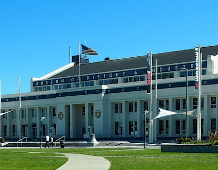 A neoclassical museum building with columns, flags, and a curved walkway; a bright day with a blue sky and people around the entrance.