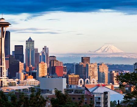 Seattle skyline with Space Needle in foreground and mountains (possibly Mount Rainier) in the distance at dusk.