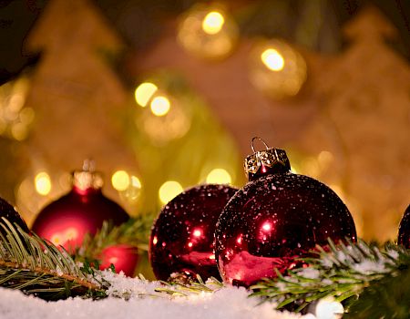 Shiny red Christmas ornaments among pine branches and snow, with warm glowing lights in the blurred background, festive and cozy.