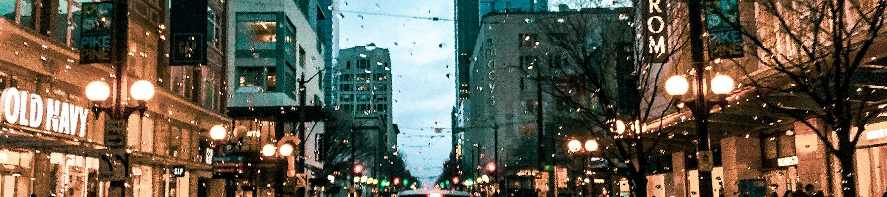 A city street at dusk with wet pavement, cars in traffic, storefronts lit up, and string lights overhead.