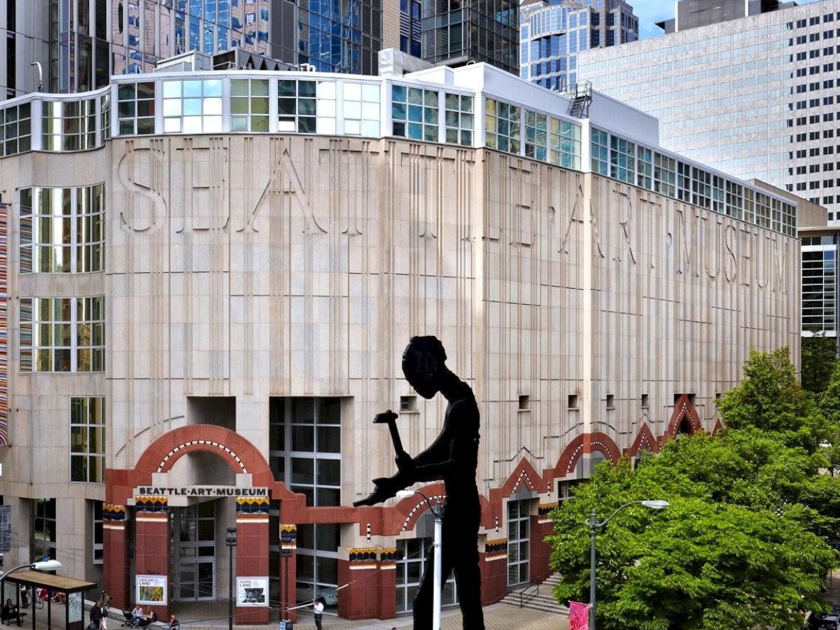 A tall silhouette of a man bending to lift or dance on a giant statue in front of a modern urban building, with trees and cars nearby.