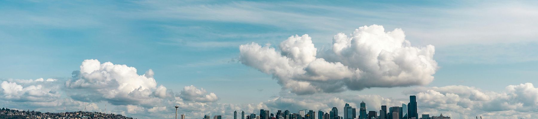 Seattle skyline across the water on a bright day with fluffy clouds overhead, calm waves, and a wide blue sky.
