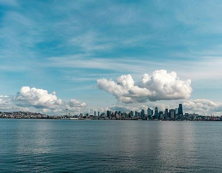 Seattle skyline across the water on a bright day with fluffy clouds overhead, calm waves, and a wide blue sky.