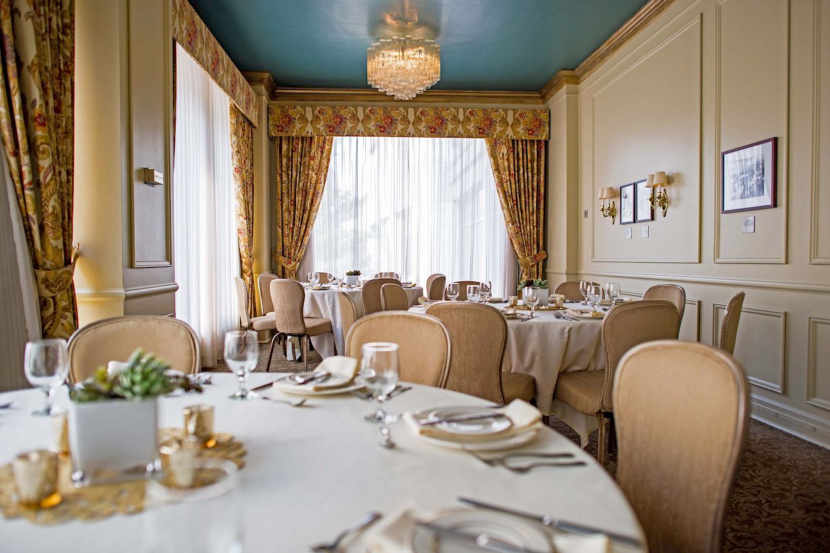 A refined dining room with round tables, cream chairs, soft gold accents, elegant drapery, and a crystal chandelier, ready for a formal meal.